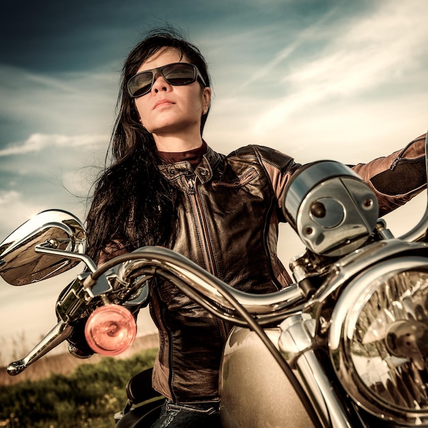 Garota motociclista em uma jaqueta de couro em uma motocicleta, olhando o pôr do sol.