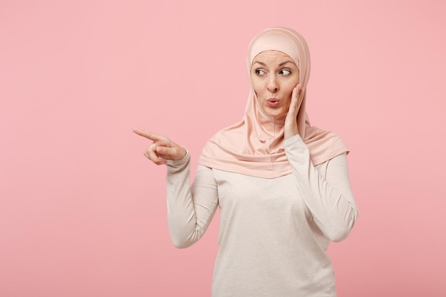 Garota muçulmana árabe em roupas leves hijab posando isolado no fundo rosa. Conceito de estilo de vida do Islã religioso de pessoas. Mock up espaço de cópia. Conta segredo com gesto de mão apontando o dedo indicador de lado.