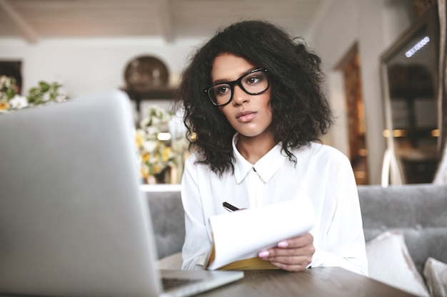 Garota muito afro-americana de óculos, trabalhando no laptop no restaurante. jovem senhora com cabelo escuro encaracolado sentada num café com o caderno na mão