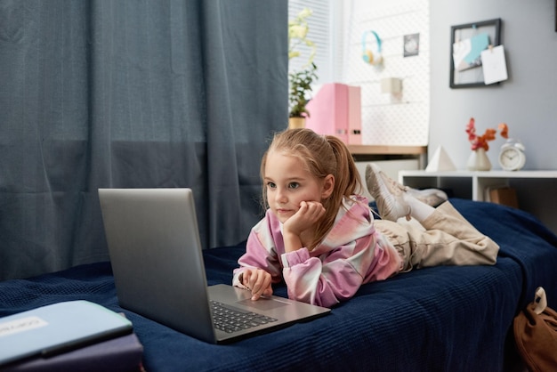 Garota muito elementar séria em roupa casual, deitada na cama e usando internet no laptop