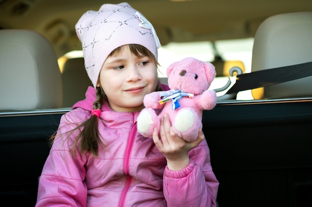 Garota muito feliz criança brincando com um urso de pelúcia rosa brinquedo sentado em uma mala do carro.