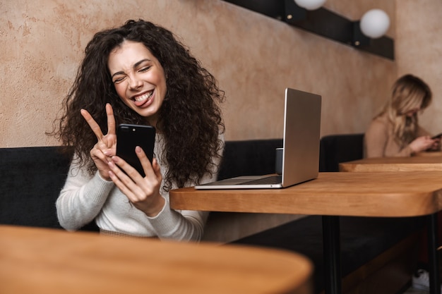 Foto garota muito feliz sentada em um café usando um laptop e um telefone celular, mostrando paz