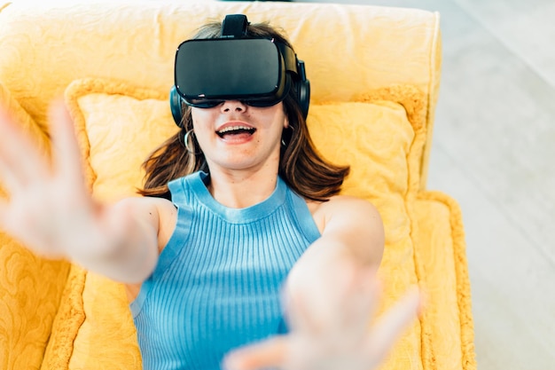 Garota muito surpresa com óculos de realidade virtual, deitada no sofá, levantando os braços no ar