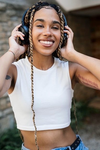 Garota multirracial com longas tranças loiras ouvindo música na rua com fones de ouvido e sorrindo para a câmera