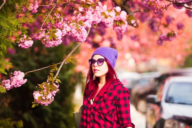 Garota na moda com longos cabelos coloridos posando perto de filial de sakura enquanto a cidade caminha. Cidade de primavera blured em um fundo.