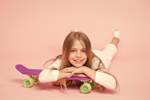 Garota na moda Garoto adorável criança cabelos longos adora andar de penny board Andar de penny board e fazer truques Garota gosta de andar de skate Estilo de vida ativo Garota se divertindo com penny board fundo rosa