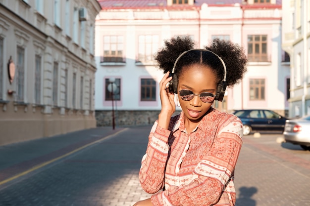 Garota negra afro-americana tem um passeio na cidade à noite e ouvindo música em fones de ouvido. Garota de óculos de sol no verão sorri e olhando no pôr do sol.