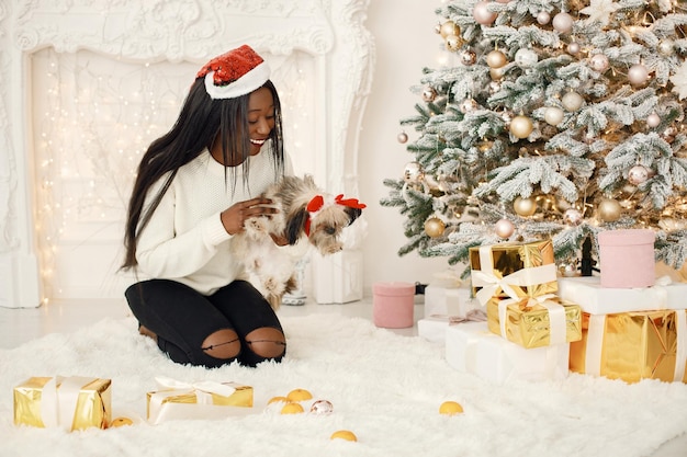 Garota negra com chapéu de Papai Noel sentada perto da árvore de Natal segurando um cachorrinho