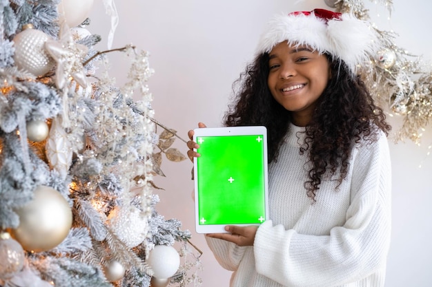 Garota negra sorridente com chapéu de Papai Noel mostrando tablet com tela verde segurando o gadget croma chave perto da árvore de Natal anunciando vendas e presentes de aplicativos de produtos de Natal