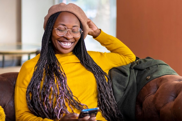 Garota negra sorrindo com dentes perfeitos sentada no sofá com o celular na mão Conceito de sorriso