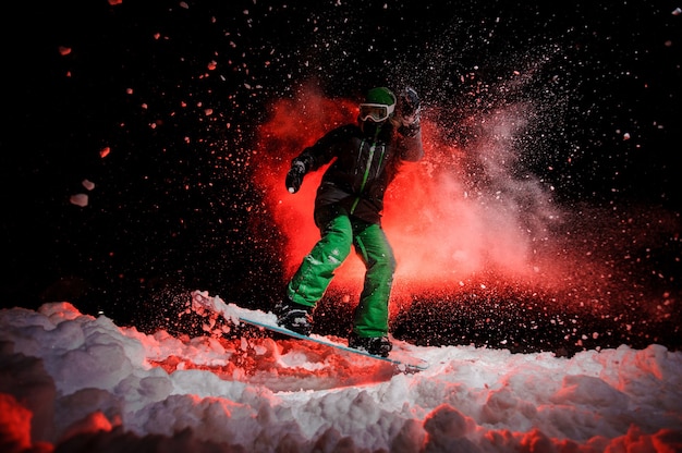 Garota no snowboard vestida com uma roupa esportiva verde, pulando na neve à noite sob a luz vermelha