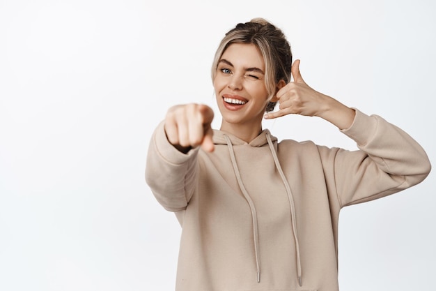 Garota piscando sorridente mostrando telefone celular me chame de gesto apontando o dedo para a câmera convidando você em pé no capuz contra fundo branco