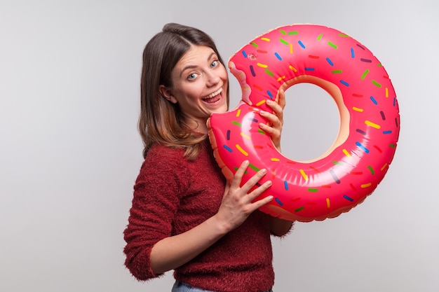 Garota positiva mordendo donut inflável grande, fingindo comer anel de borracha, se divertindo nas férias