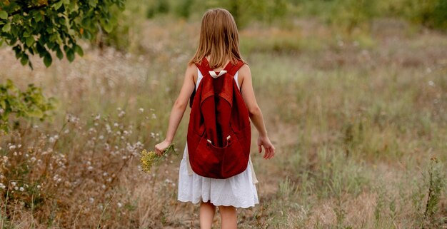 Garota pré-adolescente com mochila andando no campo num dia de verão sozinho. Criança linda na escola na natureza
