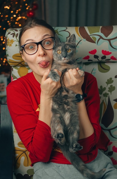 Garota relaxante segurando um gato doméstico e sentada em uma poltrona perto da árvore de Natal em uma casa aconchegante