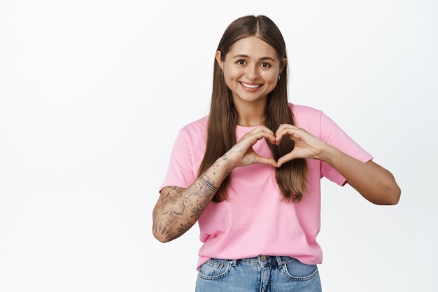 Garota romântica mostra o sinal do coração e sorri, fica na camiseta rosa com jeans em branco.