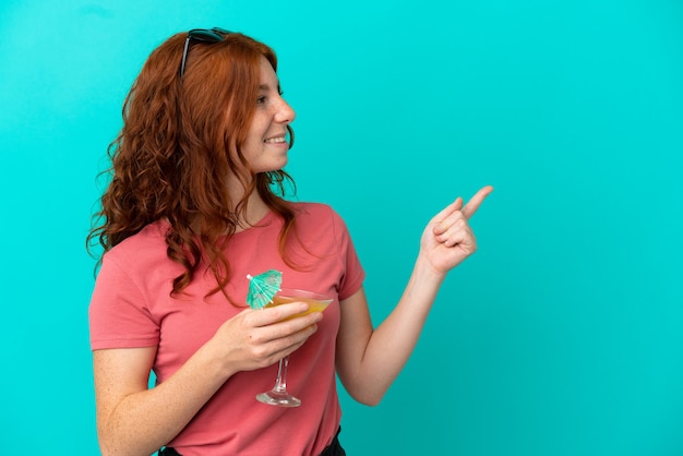 Garota ruiva adolescente segurando um coquetel isolado em um fundo azul apontando para o lado para apresentar um produto