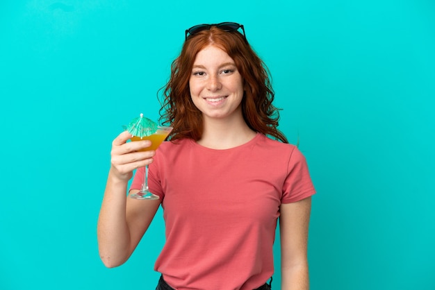 Garota ruiva adolescente segurando um coquetel isolado em um fundo azul e sorrindo muito