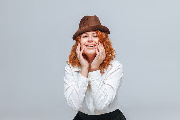 Garota ruiva alegre com um chapéu marrom em um fundo cinza e uma camisa branca