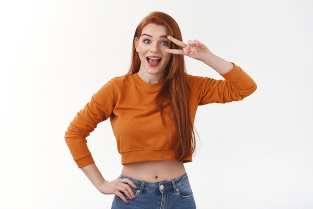 Garota ruiva bonita e boba em jeans laranja recortado segure a mão no quadril faça sinal de paz ou vitória perto do olho e sorrindo alegremente envie positividade reaja com alegria e felicidade a boas notícias