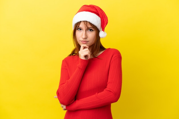 Garota ruiva com chapéu de natal isolada em um fundo amarelo pensando