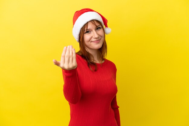 Garota ruiva com chapéu de Natal isolado em fundo amarelo, convidando para vir com a mão. Feliz que você veio