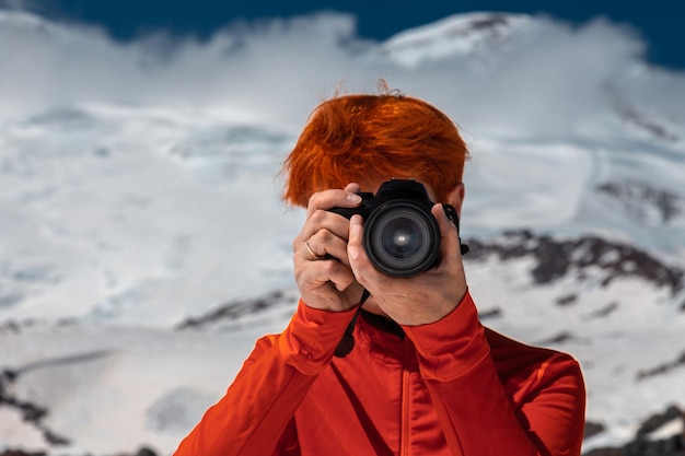Garota ruiva com um rosto escondido atrás de uma câmera voltada para o quadro contra o pano de fundo das montanhas