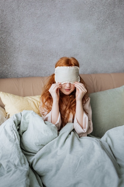 Garota ruiva com uma máscara de dormir acorda em sua cama.