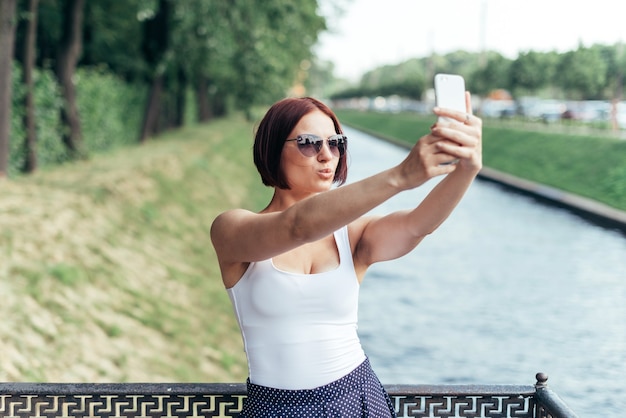 Garota ruiva em óculos de sol faz uma foto em um smartphone com um beijo.