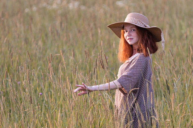 Garota ruiva emocional brincando na grama em um prado de verão ao pôr do sol em um vestido elegante