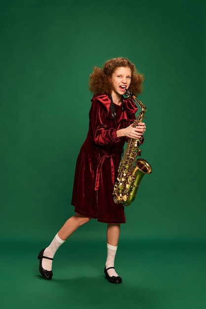 Garota ruiva encaracolada feliz em idade escolar usando vestido festivo tocando saxofone sobre verde