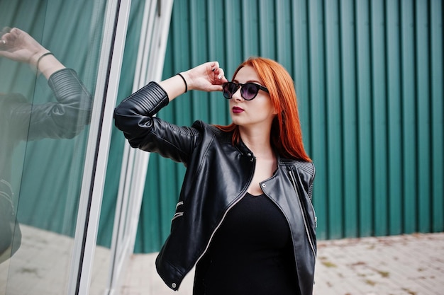 Garota ruiva estilosa em óculos de sol se veste de preto contra grandes janelas Retrato de moda de rua