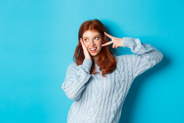 Garota ruiva feliz sorrindo, mostrando o símbolo da paz e olhando para a esquerda na promoção, vestindo um suéter contra um fundo azul