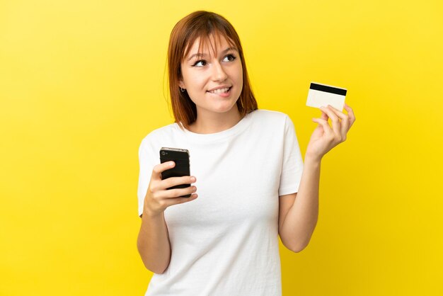 Garota ruiva isolada em fundo amarelo comprando com o celular com cartão de crédito enquanto pensava