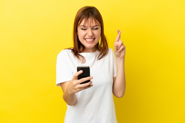 Garota ruiva isolada em fundo amarelo usando telefone celular com dedos cruzando