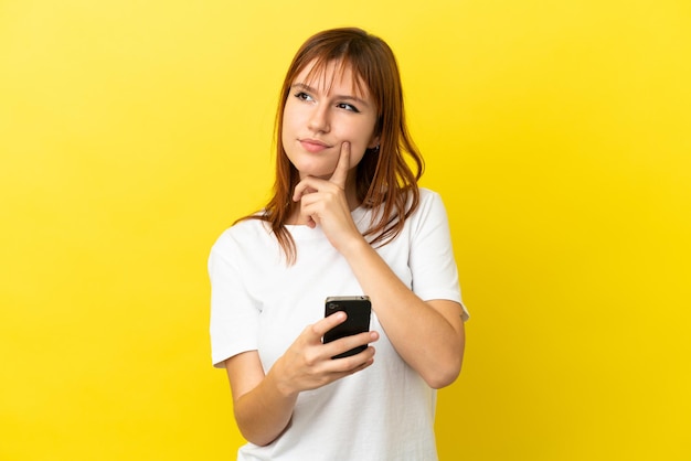 Garota ruiva isolada em um fundo amarelo usando telefone celular e pensando