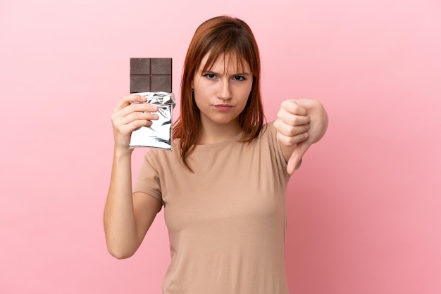 Garota ruiva isolada em um fundo rosa tomando uma pastilha de chocolate e fazendo um sinal ruim