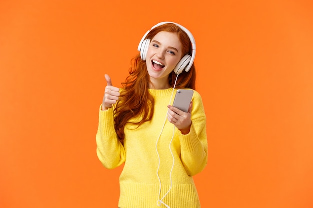 garota ruiva receber presente de Natal novos fones de ouvido legais, ouvindo música em fones de ouvido e mostrando o polegar para cima, segurando o smartphone