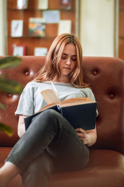 Garota ruiva sentada em um sofá na biblioteca pública com a perna em cima da outra lendo um livro com interesse.