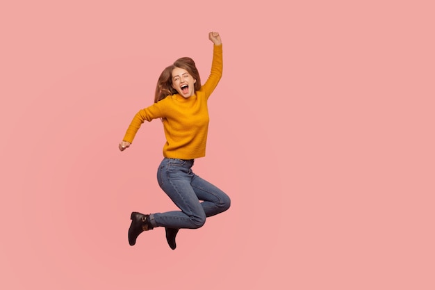 Garota ruiva vívida e energética no suéter e jeans pulando no ar ou voando gritando Sim, eu fiz isso comemorando o sucesso da vitória cheio de entusiasmo estúdio interno tiro fundo rosa de corpo inteiro