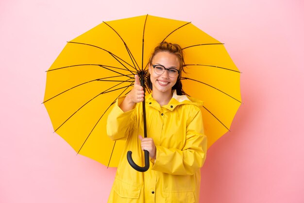 Garota russa adolescente com casaco à prova de chuva e guarda-chuva isolado em um fundo rosa apertando as mãos para fechar um bom negócio