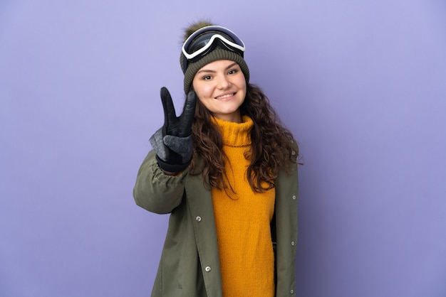 Garota russa adolescente com óculos de snowboard isolados na parede roxa sorrindo e mostrando sinal de vitória