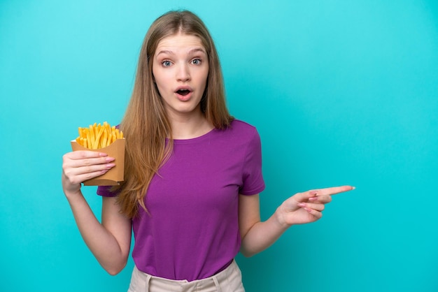 Garota russa adolescente pegando batatas fritas isoladas em fundo azul surpresa e apontando o lado