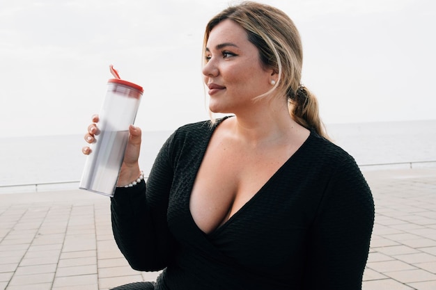 Garota russa com sardas plussize bebendo água depois de se exercitar em frente à praia