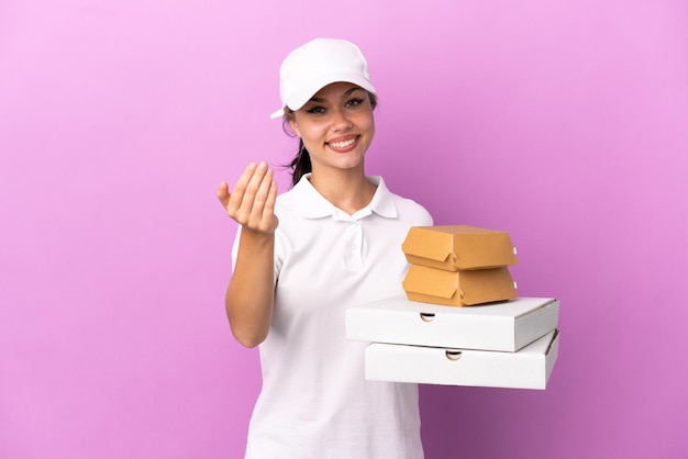 Garota russa de entrega de pizza com uniforme de trabalho pegando caixas de pizza e hambúrgueres isolados em fundo roxo convidando para vir com a mão Feliz por você ter vindo