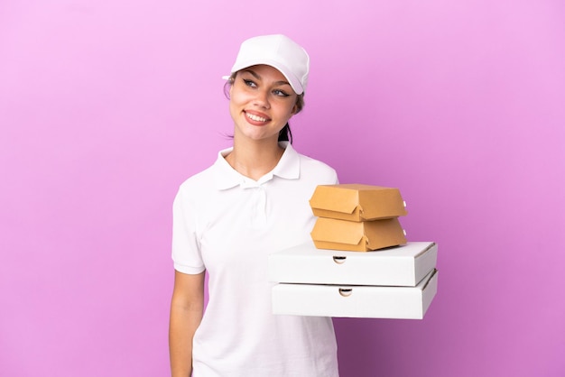 Garota russa entregadora de pizza com uniforme de trabalho pegando caixas de pizza e hambúrgueres isolados no fundo roxo olhando para o lado e sorrindo