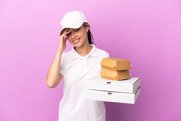 Garota russa entregadora de pizza com uniforme de trabalho pegando caixas de pizza e hambúrgueres isolados no fundo roxo rindo