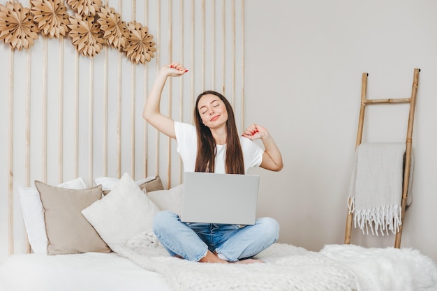 Garota se estica preguiçosamente enquanto está sentada na cama com um laptop Conceito de estilo de vida freelancer de trabalho remoto