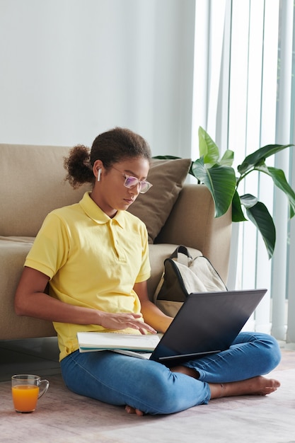 Garota séria e inteligente de raça mista em óculos, sentada com as pernas cruzadas no chão e fazendo tarefas usando um laptop