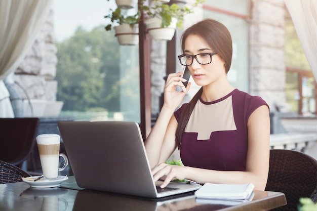 Garota séria, falando no celular e usando o laptop durante o almoço, sentado no terraço do café. Retrato do estilo de vida da jovem mulher de negócios. Conceito de tecnologia e comunicação online